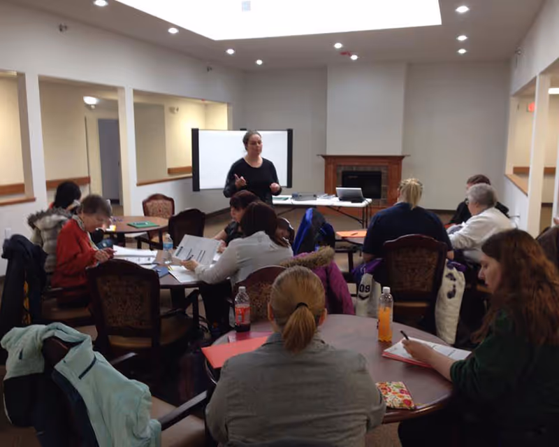 A group of people seated around tables in a room, engaged in a meeting or class. A woman stands at the front near a whiteboard and a table with a laptop and papers. The room has a fireplace and recessed lighting in the ceiling.