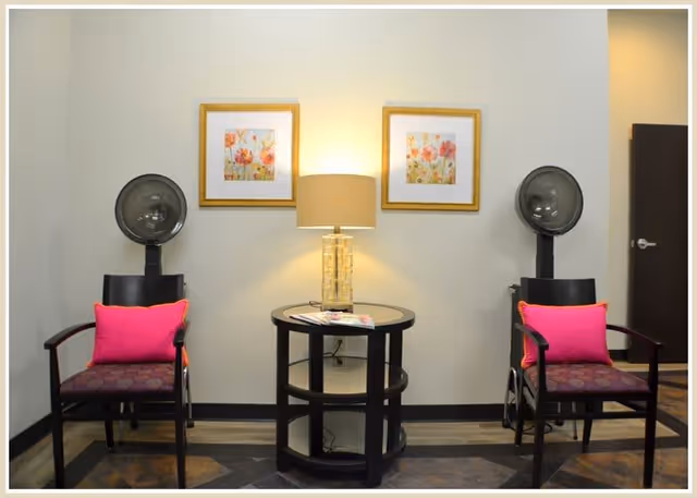 A cozy salon area with two black hair dryer chairs, each with a pink cushion. Between the chairs is a round black table with a glass lamp and some magazines. Two framed floral paintings hang on the wall above the lamp.