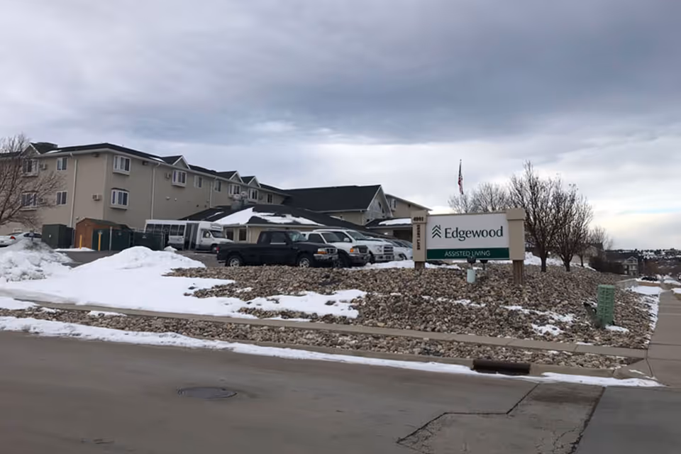 Exterior view of Edgewood Assisted Living facility in a snowy setting with several parked vehicles and a sign displaying the facility's name. The sky is overcast and there are leafless trees around the property.