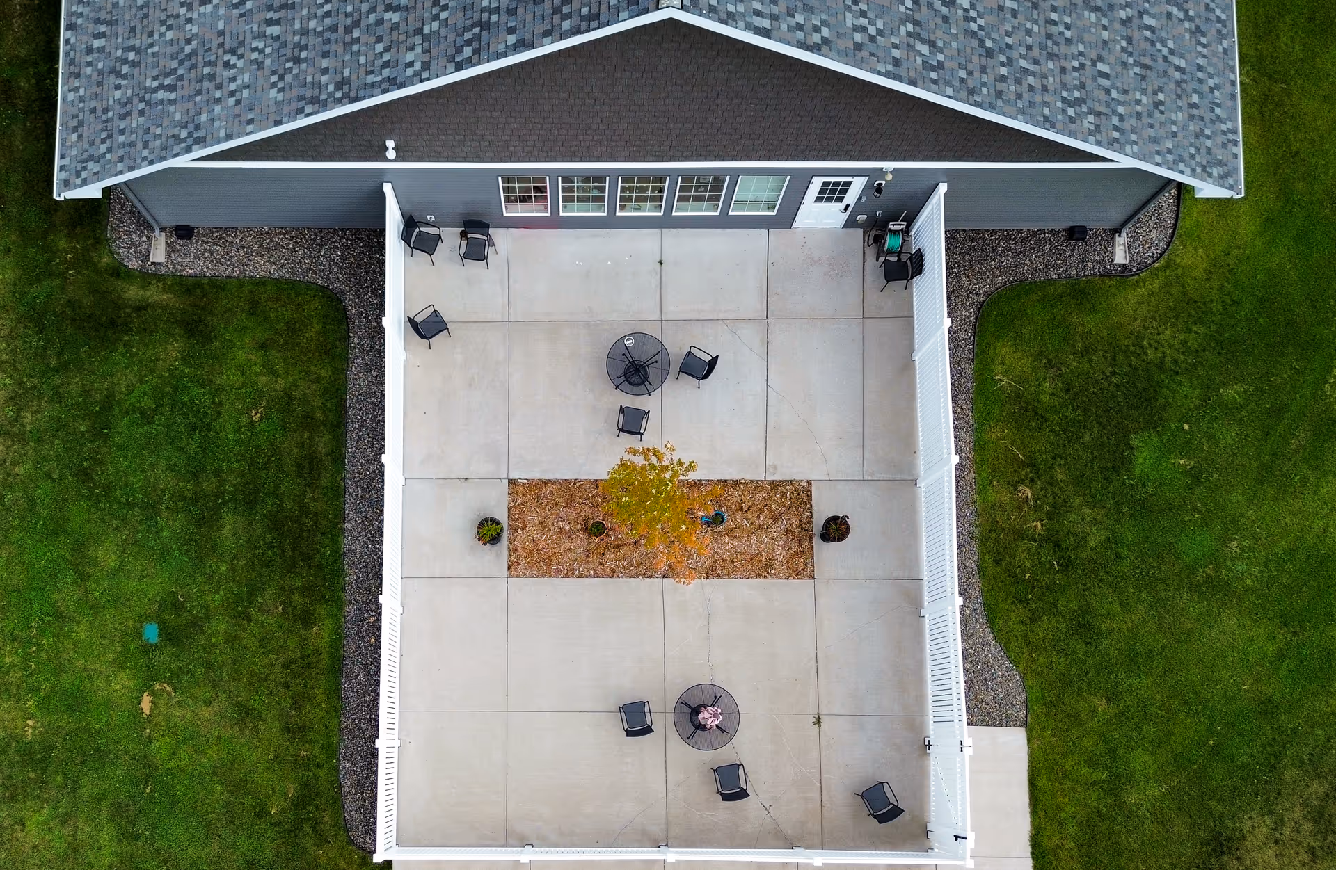 Aerial view of a fenced concrete patio area attached to a building with a shingled roof. The patio has several black chairs and two round tables, one with an umbrella. There is a rectangular garden bed with a small tree and plants in the center of the patio. Surrounding the patio is green grass and landscaping rocks along the building's foundation.
