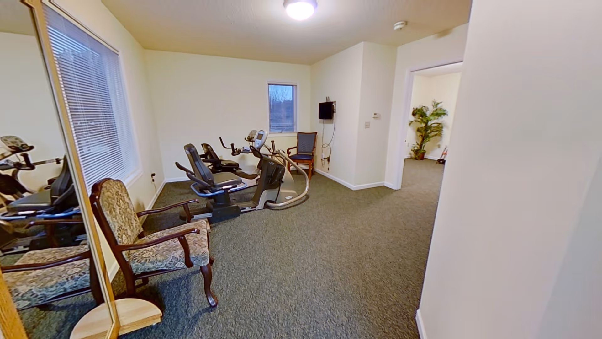 A small exercise room with two stationary recumbent bikes, a wooden chair with patterned upholstery, a wall-mounted TV, and a window with blinds. The room has beige walls and carpeted flooring, with an open doorway leading to another room with a large potted plant.