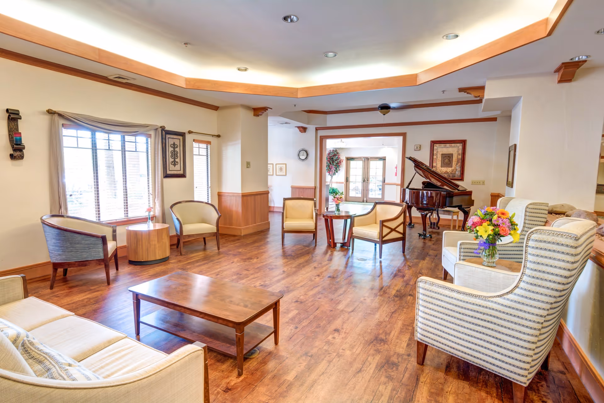 A spacious and well-lit living room area with wooden flooring, beige and striped upholstered chairs, a wooden coffee table, and side tables with flower vases. Large windows with blinds and curtains allow natural light to fill the room. A grand piano is positioned near the back wall, next to framed artwork and a clock. The ceiling features recessed lighting with wooden trim accents.