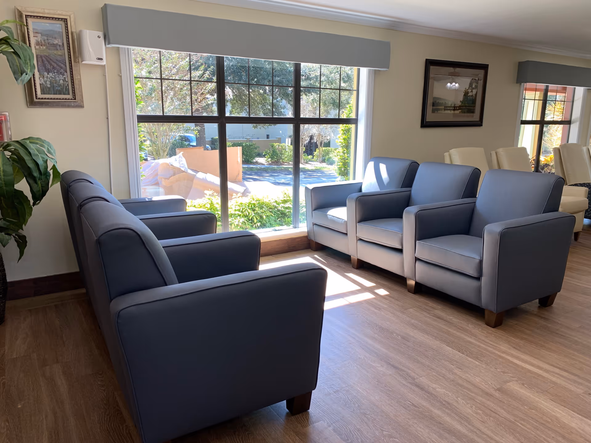 A bright and cozy seating area with four gray armchairs arranged in two pairs facing each other near a large window that lets in natural light. The window offers a view of greenery and outdoor seating. The room has wooden flooring, framed artwork on the walls, and additional beige chairs visible in the background.