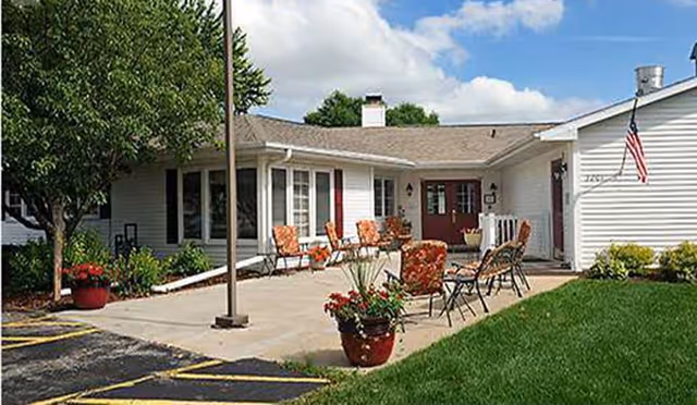 Front entrance patio of a senior living facility with outdoor chairs, potted plants, an American flag, and the building facade.