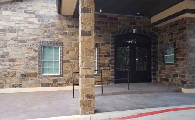 Entrance area of a building with stone walls, two windows with white frames, a stone pillar in the center, and a double black door under a covered porch.