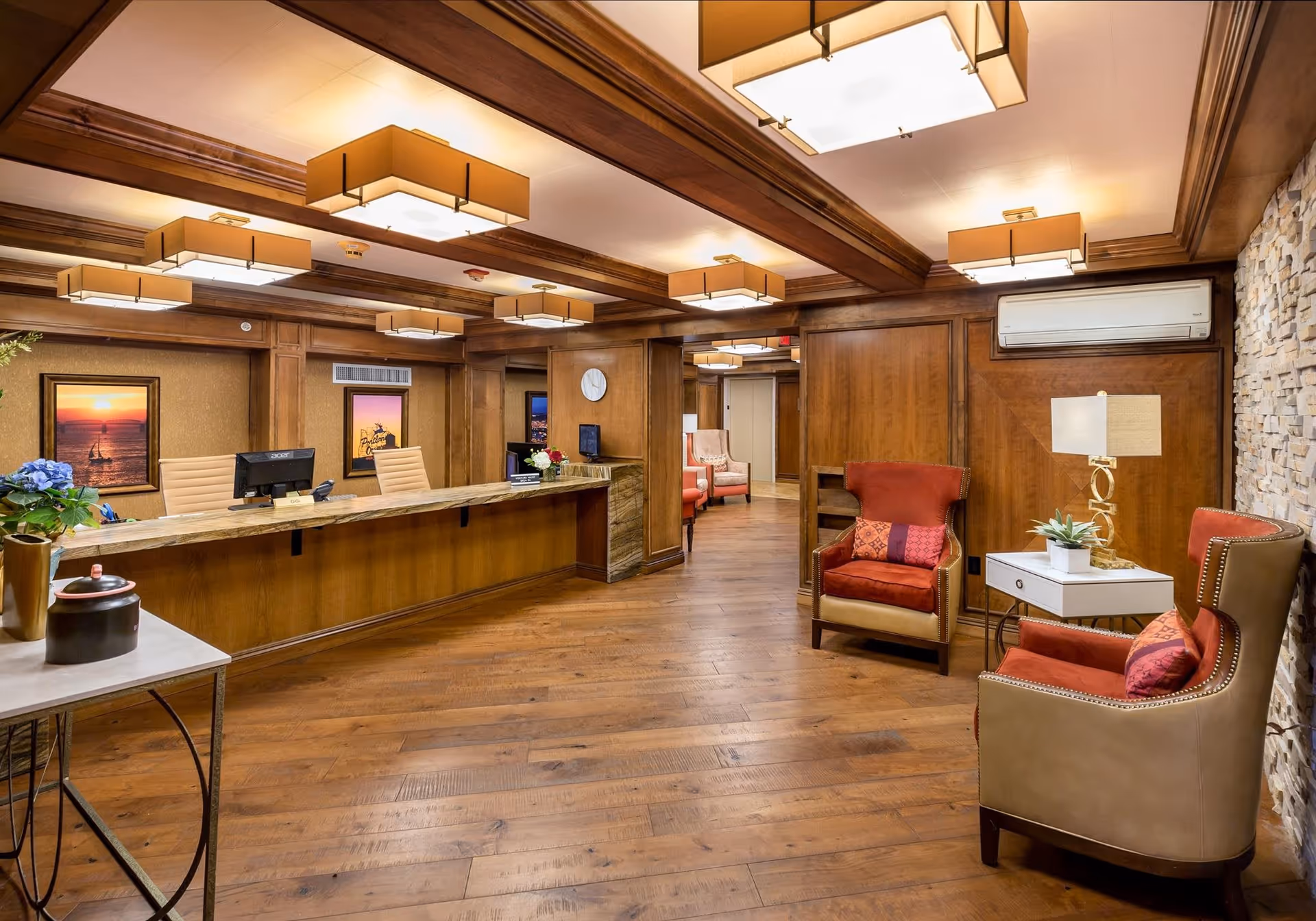 Reception area of Calaroga Terrace featuring a long wooden front desk with two chairs and computer monitors. The room has wooden floors, wood-paneled walls, and a stone accent wall. There are several modern rectangular ceiling lights, two red cushioned armchairs with decorative pillows, a small white side table with a lamp and plant, and framed sunset artwork on the walls.