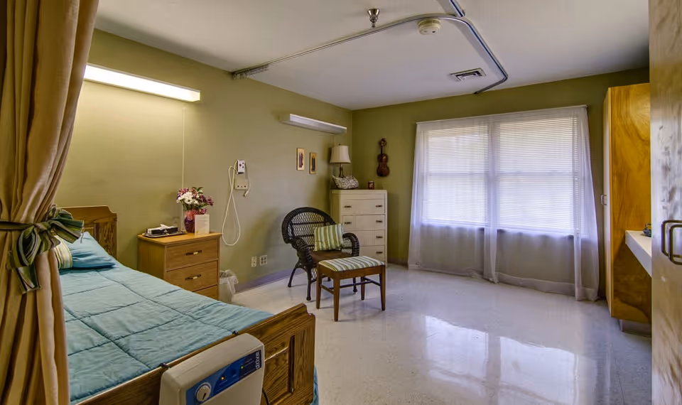 A simple, clean bedroom in a senior living facility with a single bed covered in a blue blanket, a wooden nightstand with a vase of flowers, a wicker chair with a striped cushion, a wooden dresser, and a window with sheer white curtains letting in natural light.