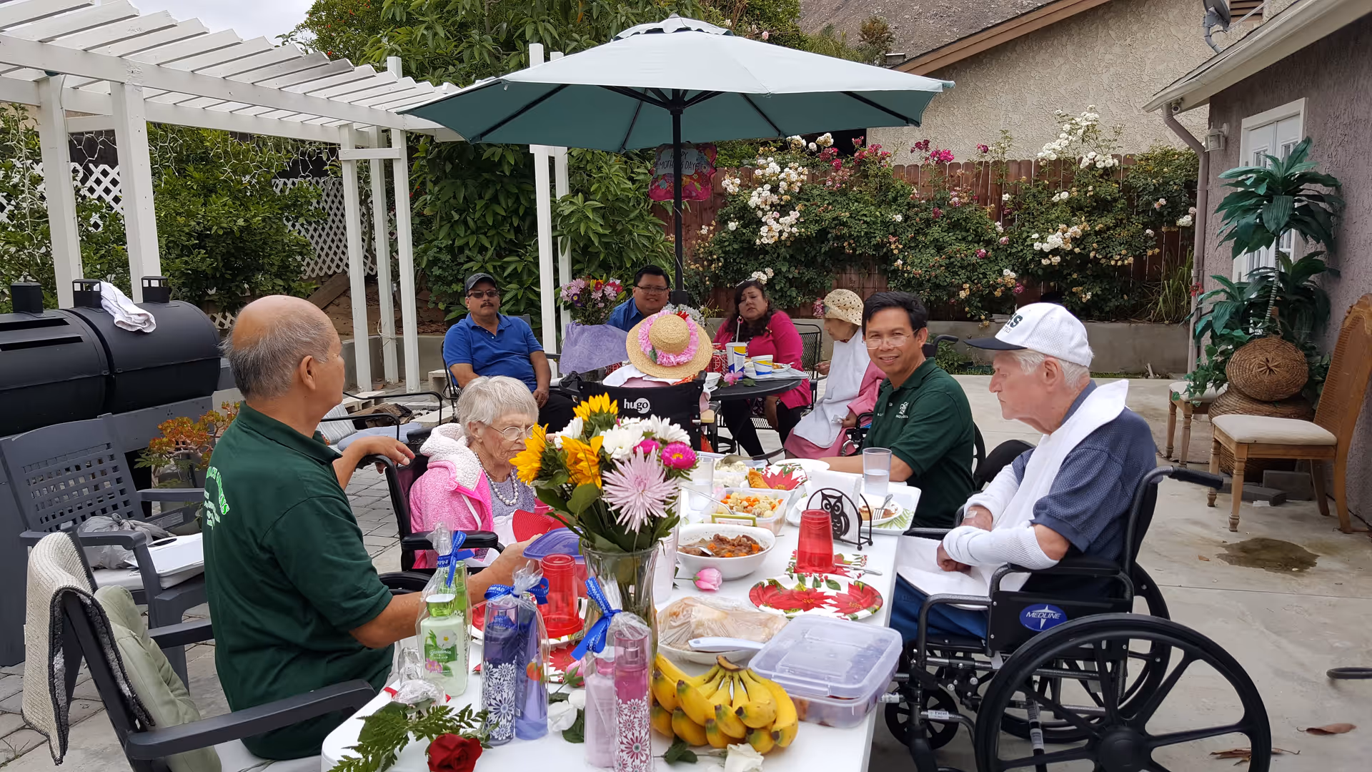 A group of elderly people and caregivers sitting around a long outdoor table covered with food, flowers, and drinks. The setting is a backyard patio with a large umbrella, greenery, and a barbecue grill in the background. Some individuals are in wheelchairs, and everyone appears to be enjoying a social gathering.