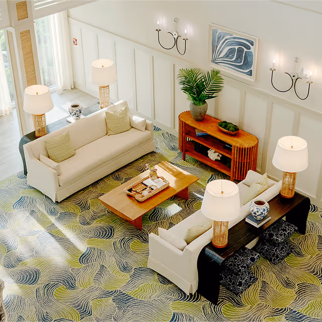 A bright and airy living room with two cream-colored sofas facing each other across a wooden coffee table. The room features a patterned carpet in shades of green and blue, a wooden console table with a large potted plant and decorative items, wall sconces with candles, and large windows letting in natural light.