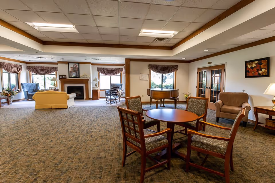 A spacious living room area in an assisted living facility featuring a round wooden table with four cushioned chairs in the foreground. In the background, there is a beige sofa, two blue armchairs, a rocking chair, a fireplace with a framed picture above it, a grand piano, and a brown armchair next to a side table with a lamp. Large windows with valances allow natural light to fill the room.