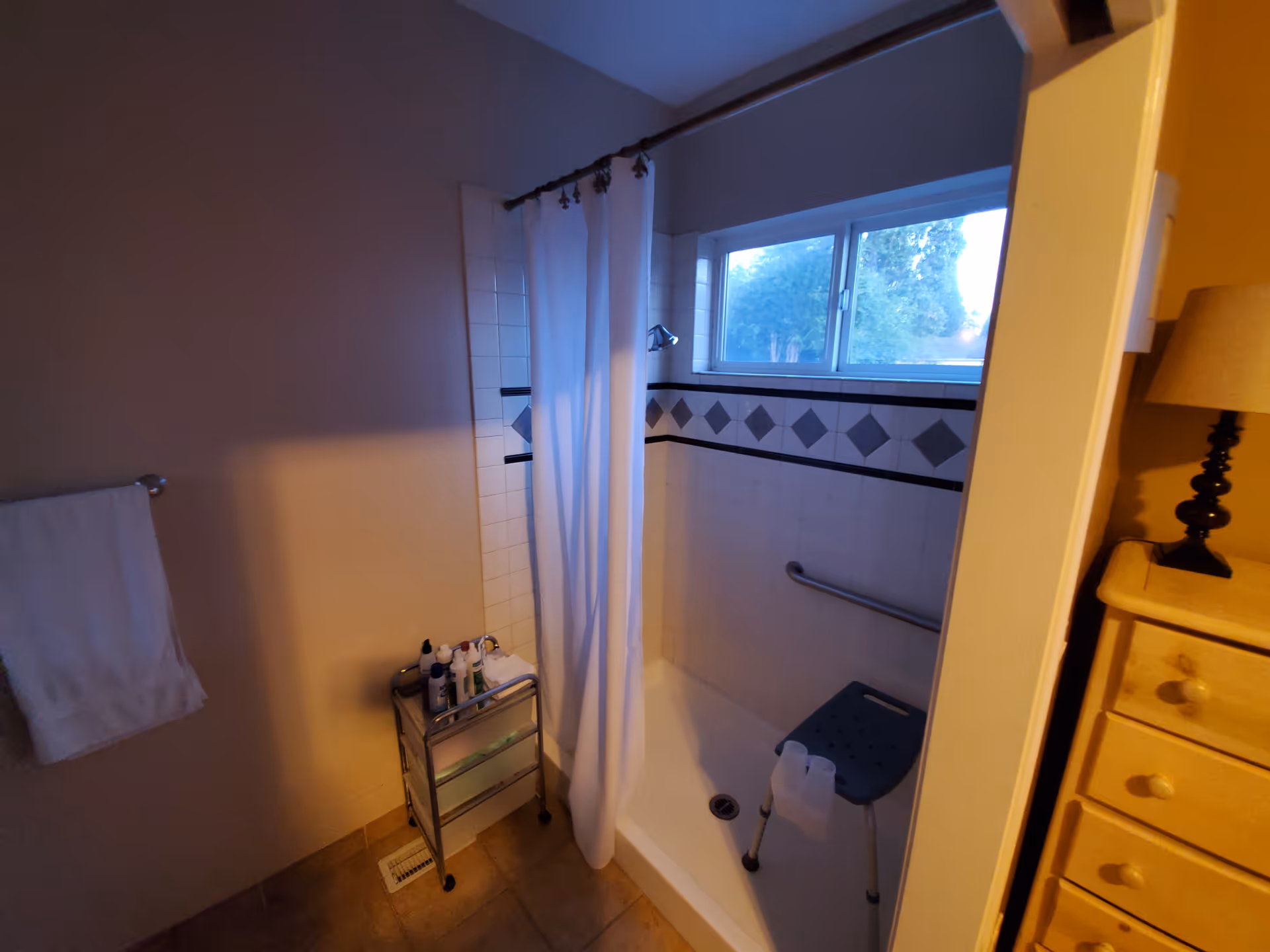 A bathroom shower area with a white shower curtain, a shower chair, and a grab bar on the tiled wall. There is a window above the shower and a small metal cart with toiletries next to the shower. A towel hangs on a rack on the left wall, and part of a wooden dresser with a lamp is visible on the right.