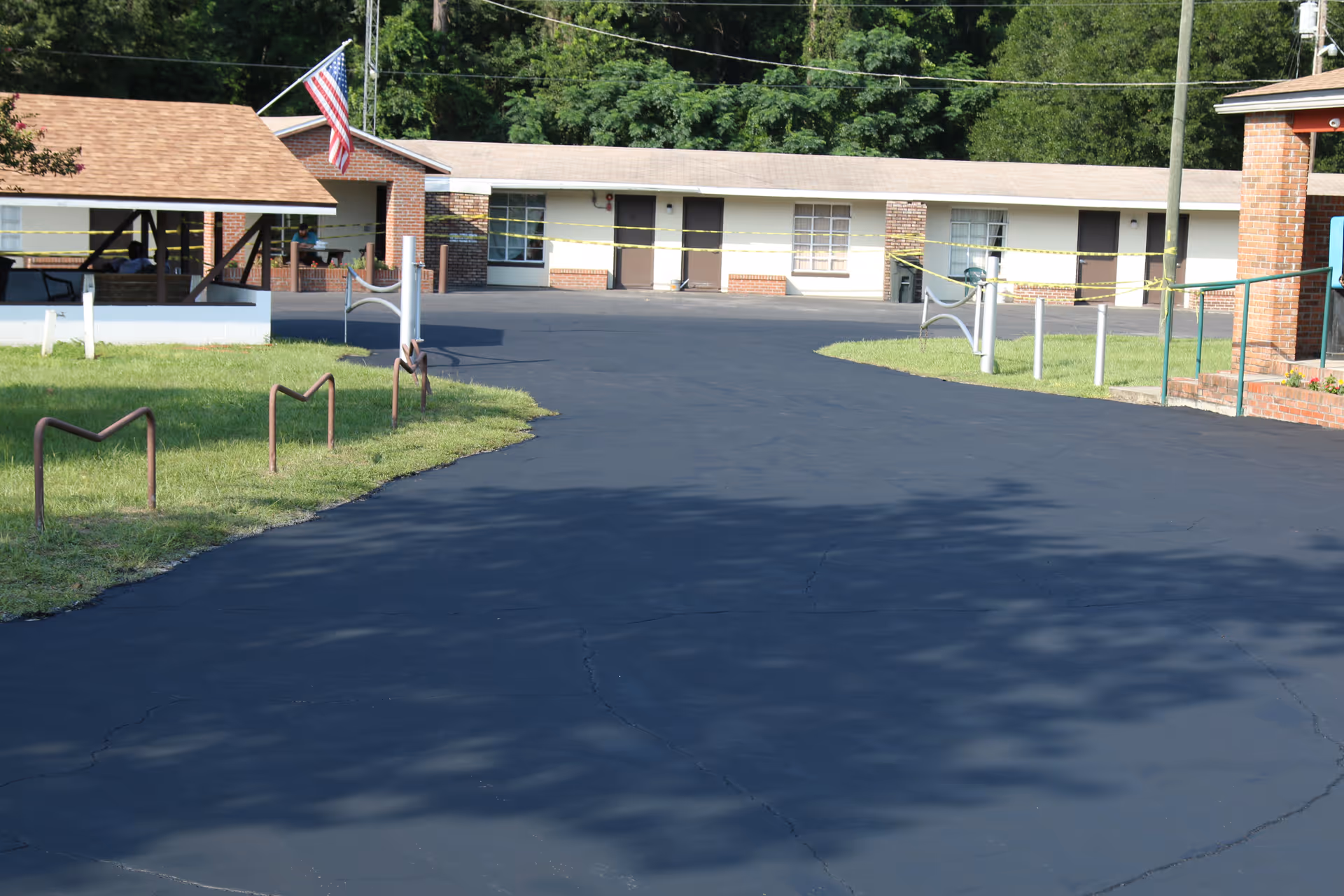 Paved driveway and grassy area leading to a single-story brick and stucco building with an American flag and rooms visible, sections cordoned off with yellow tape.