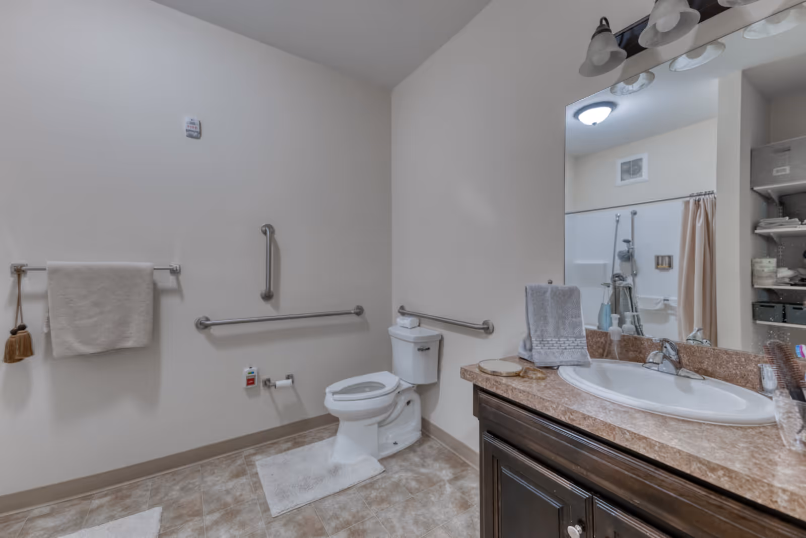 A clean and accessible bathroom with a toilet equipped with grab bars on the walls, a towel rack with a towel and brush hanging, a sink with a large mirror above it, and shelves with toiletries and towels. The floor is tiled and there are white bath mats on the floor.