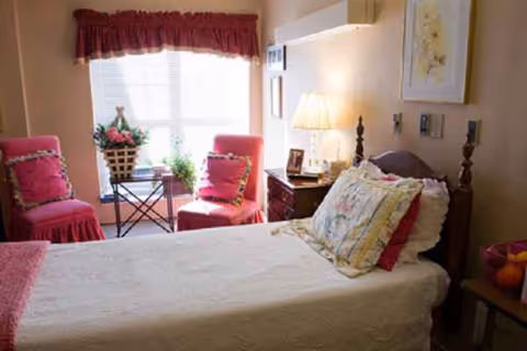 A bedroom in a senior living facility with a bed, two pink chairs, and a window.