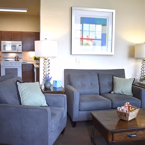 A cozy living room area with a gray armchair and matching sofa, each with light green pillows. There are two side tables with lamps featuring spiral metal bases. A wooden coffee table with a basket of snacks is in front of the sofa. In the background, a kitchen with wooden cabinets, a microwave, and a stove is visible. A framed abstract artwork hangs on the wall above the sofa.