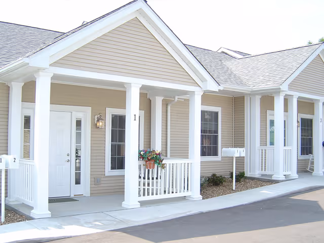 Exterior view of a beige senior living facility with white trim and columns supporting a covered porch. There are multiple units with white doors and windows, each unit numbered. A small flower pot hangs on the railing of one porch, and mailboxes are visible near the sidewalk.