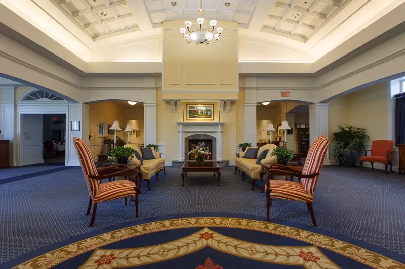 A spacious and elegant living room area with a high coffered ceiling and chandelier. The room features a fireplace with a painting above it, two beige sofas with cushions facing each other, two striped armchairs, a wooden coffee table with a floral arrangement, and several potted plants. The carpet has a decorative pattern near the foreground, and there are archways leading to other rooms.