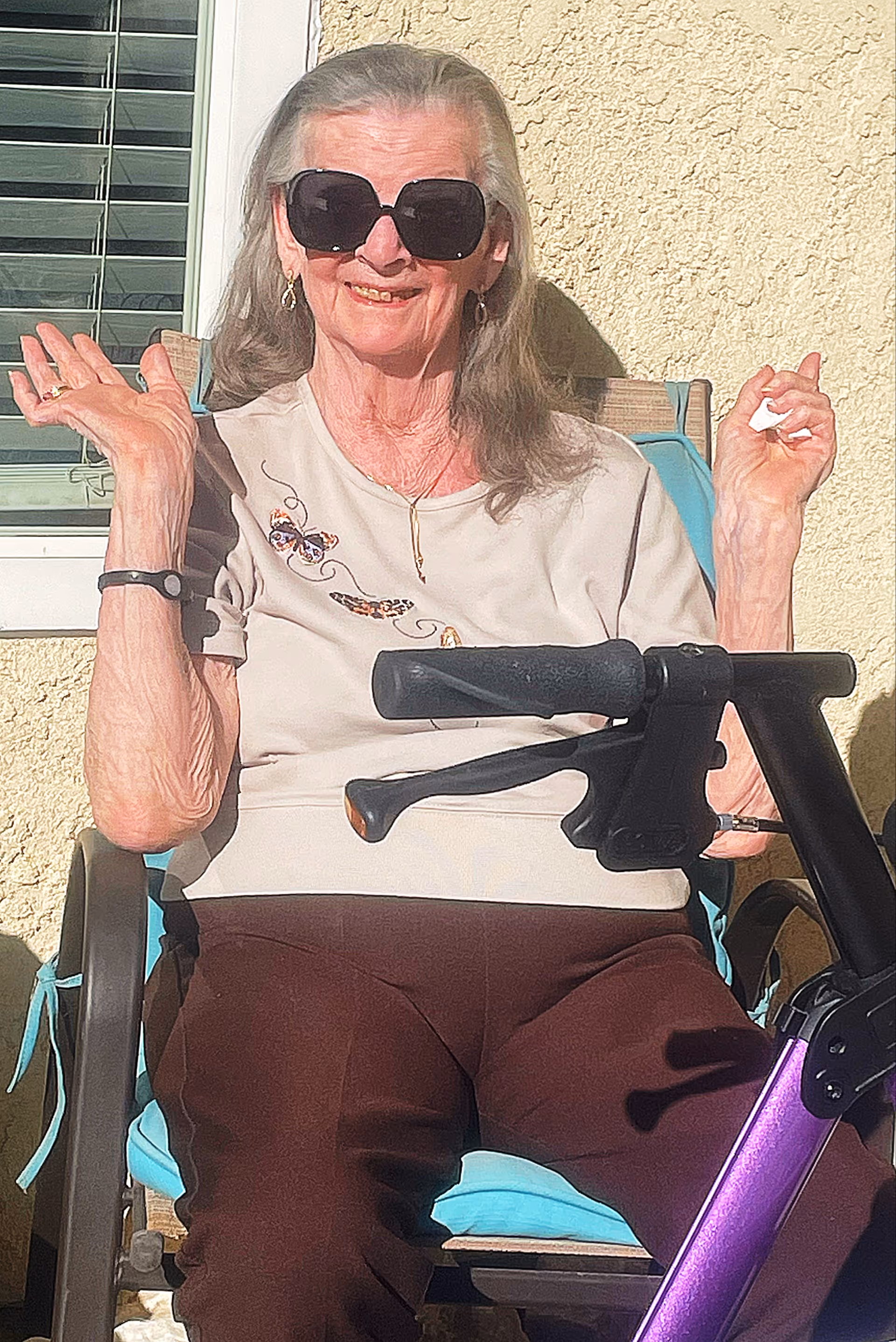 An elderly woman with long gray hair wearing large black sunglasses, a beige shirt with butterfly designs, and brown pants is sitting on a blue cushioned chair outdoors. She is smiling and waving with both hands. In front of her is a purple mobility scooter handlebar. The background shows a textured yellow wall and a window with white blinds.