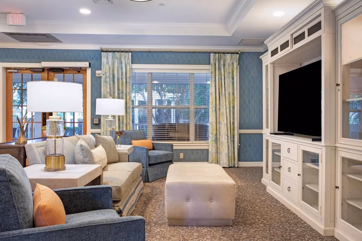 Cozy living room with sofas and armchairs around a tufted ottoman, lamps, a large TV in a white built-in cabinet, and windows with patterned curtains.