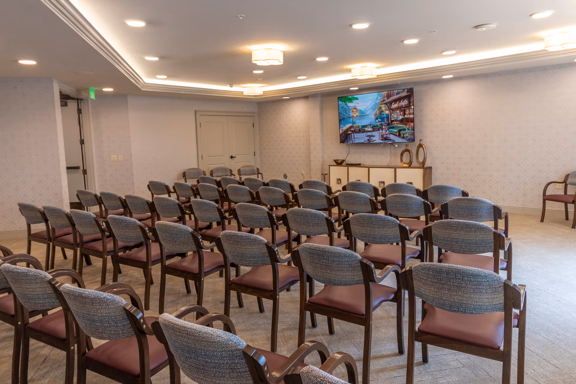 Empty meeting room with rows of upholstered chairs facing a wall-mounted TV and a cabinet.