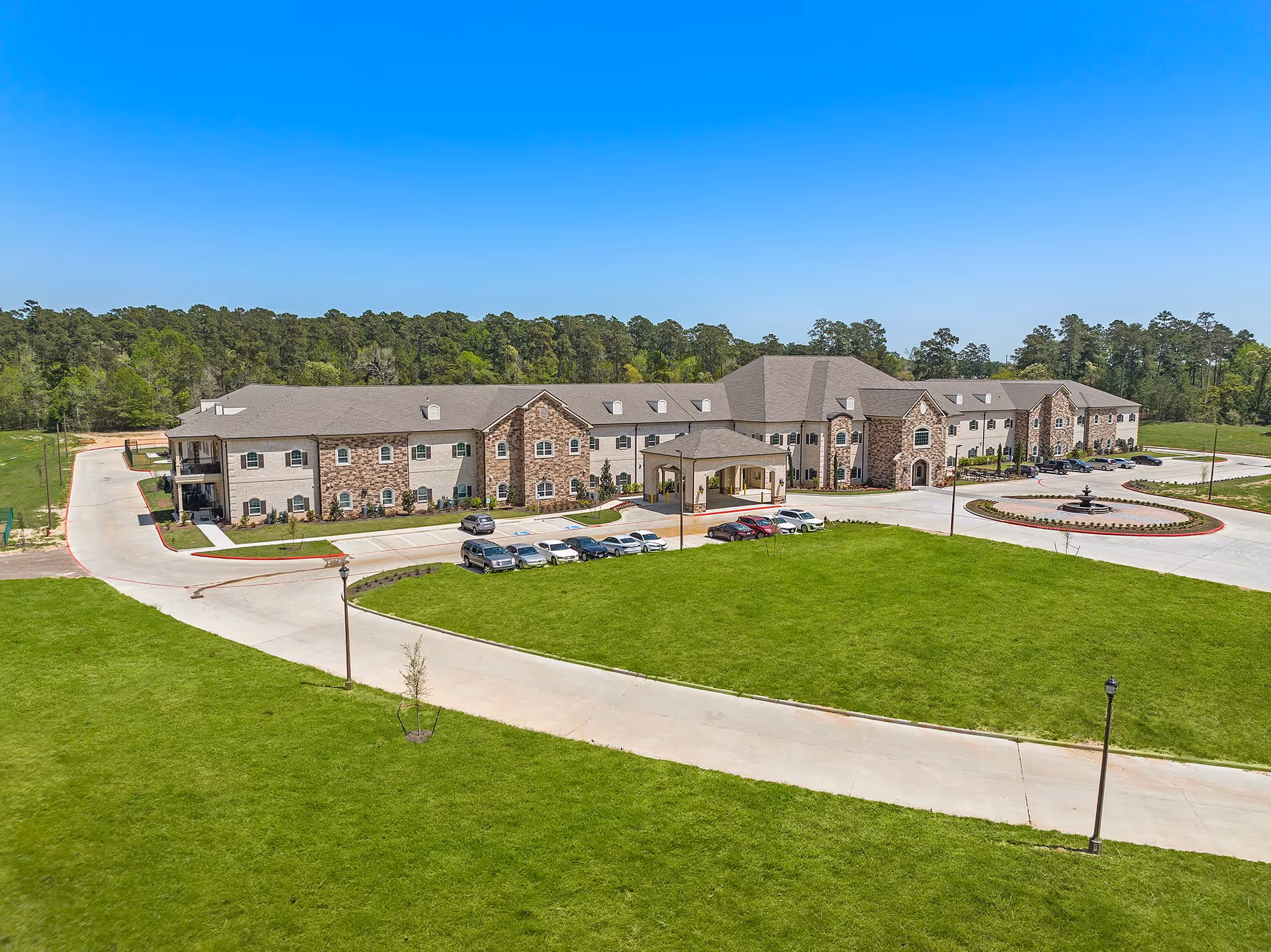 A large senior living facility building named Worthington Manor with a stone and brick facade, surrounded by a well-maintained green lawn and a circular driveway with parked cars under a clear blue sky.