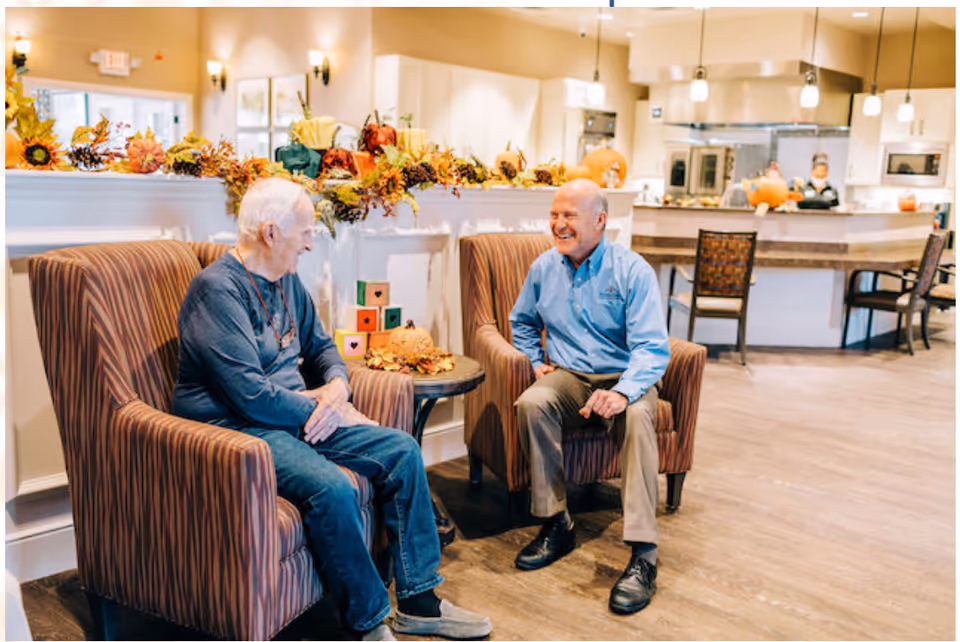 Two elderly men sitting and chatting in comfortable armchairs in a warmly decorated common area with autumn-themed decorations including pumpkins and fall leaves. In the background, a kitchen area with a staff member is visible.