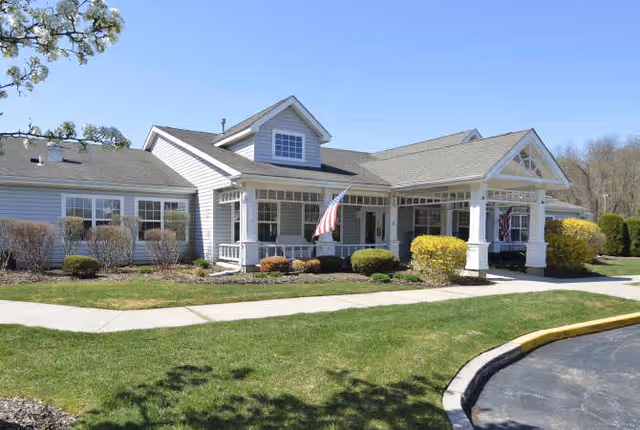 Exterior view of a single-story senior living facility building with white siding, a covered entrance supported by white columns, an American flag displayed near the entrance, manicured bushes, and a clear blue sky.