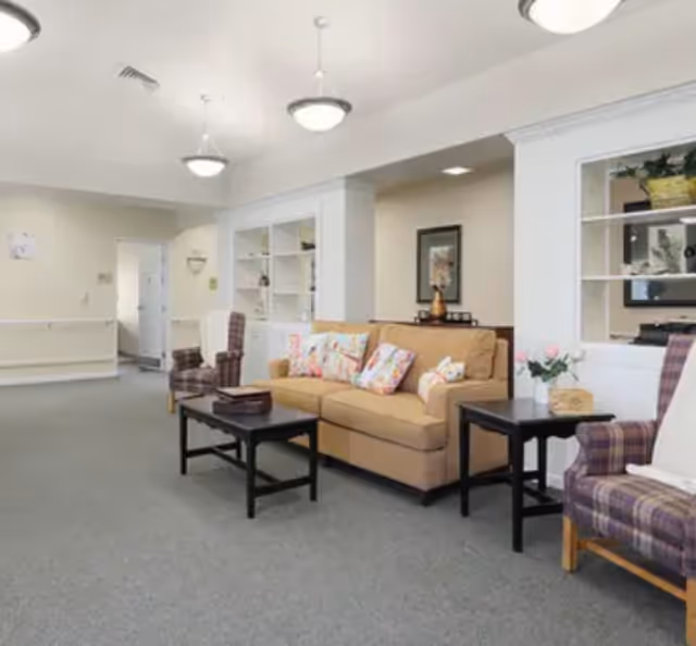 A cozy living room area in a senior living facility with a beige sofa adorned with floral pillows, two plaid armchairs, two black coffee tables, built-in white shelves with decorative items, and soft overhead lighting.
