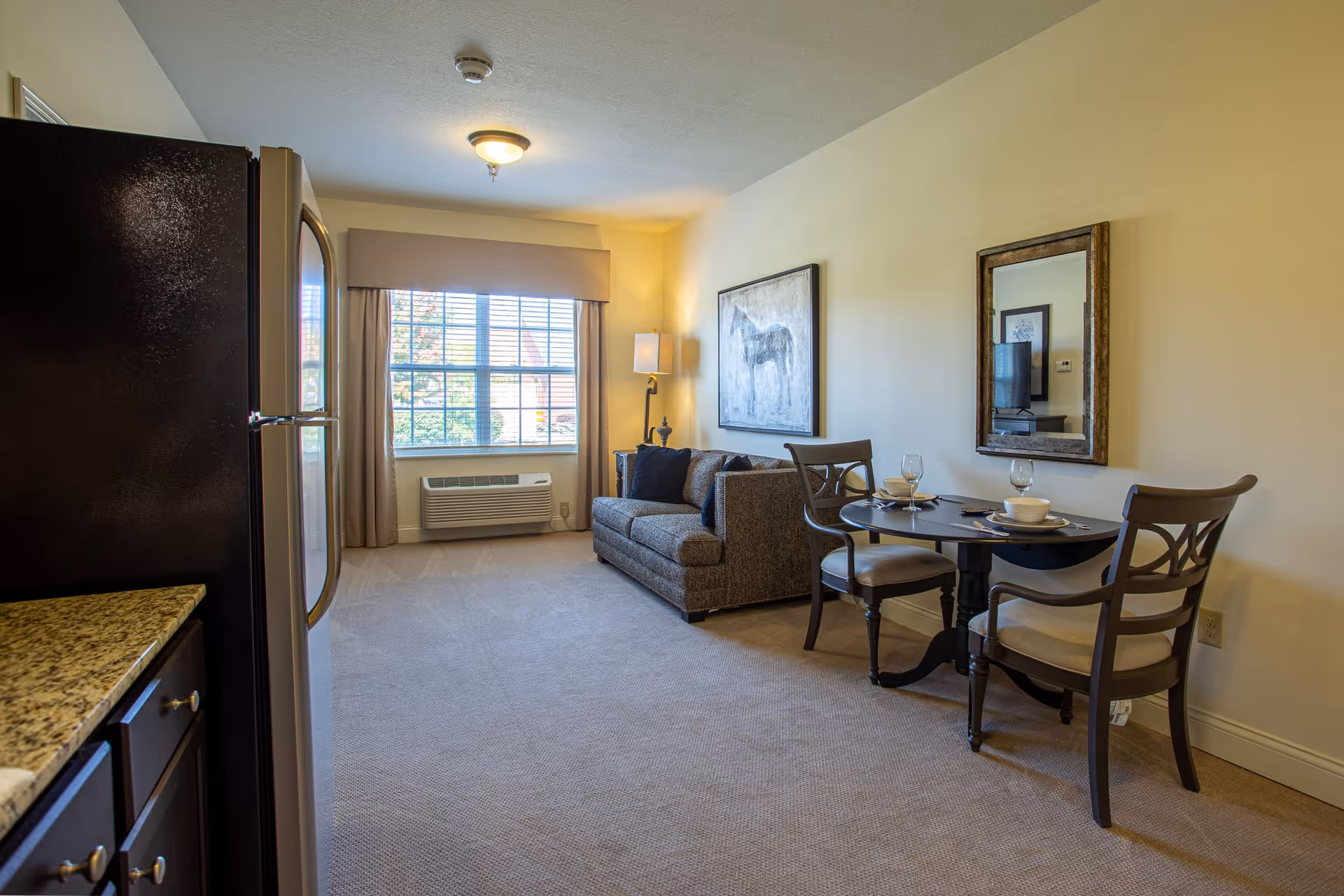 A cozy living area in a senior living facility featuring a small round dining table set for two with chairs, a gray upholstered sofa with dark cushions, a floor lamp, and a large window with beige curtains letting in natural light. The room has beige walls, carpeted floor, and a kitchen area with granite countertops and a refrigerator visible on the left side.