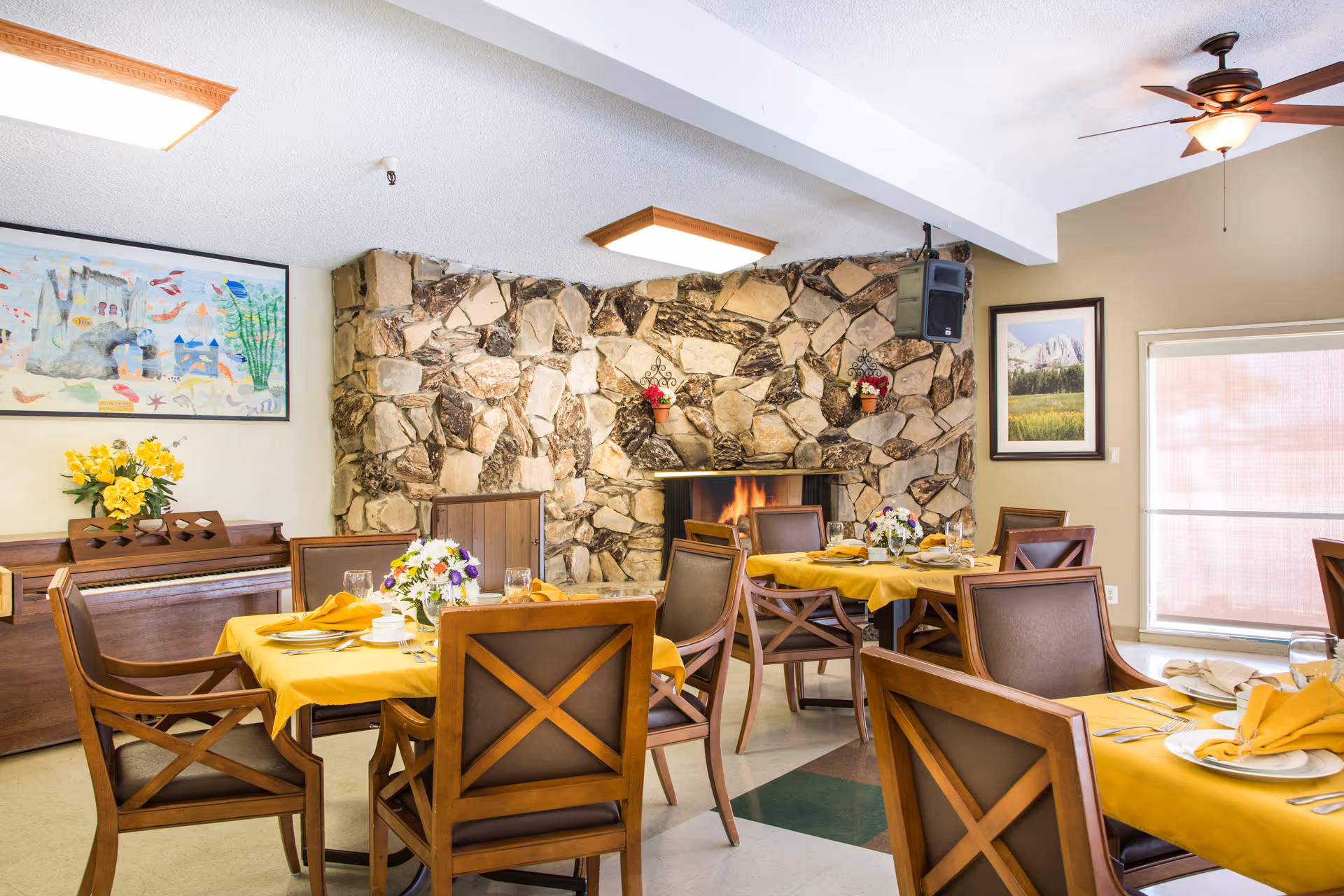 Dining room with several wooden tables covered with yellow tablecloths, set with plates, glasses, and napkins. Wooden chairs surround the tables. A stone fireplace with a fire burning is visible in the background, along with a piano, floral arrangements, and framed artwork on the walls.
