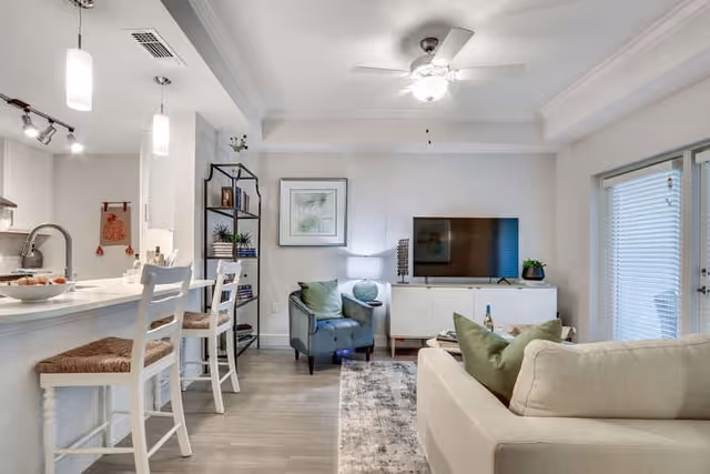 A bright and modern living room with a white sofa adorned with green pillows, a gray armchair, a white media console with a flat-screen TV, and a black metal shelving unit with decorative items. The room features light wood flooring, a ceiling fan with a light, and a kitchen counter with two white bar stools. Large windows with blinds allow natural light to enter the space.