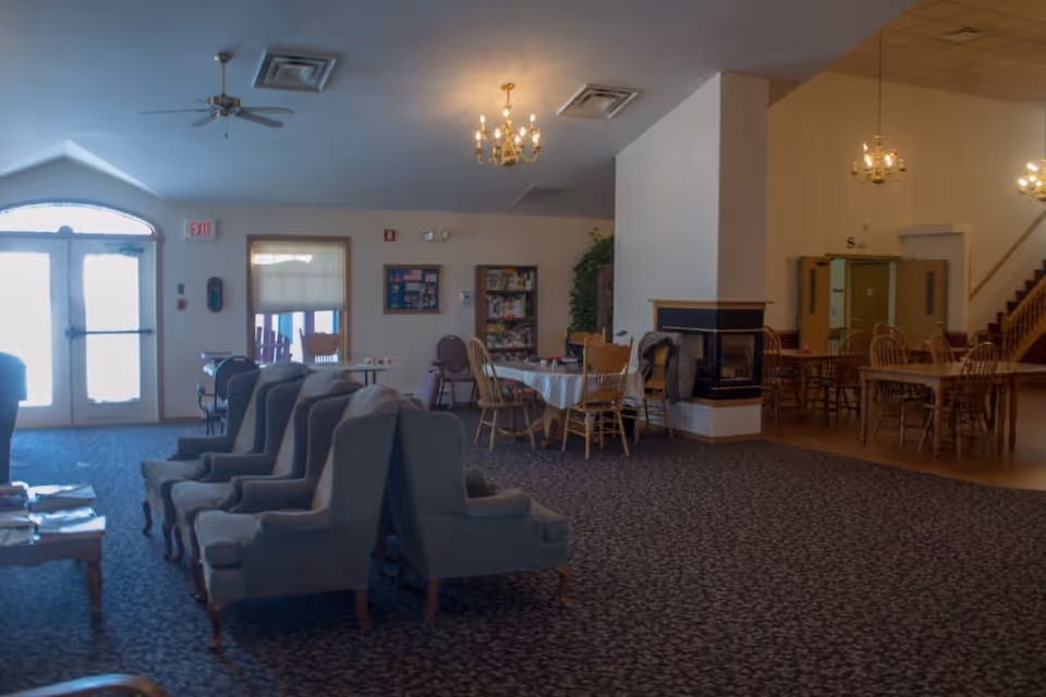 Interior view of a senior living facility common area with multiple seating arrangements including armchairs and wooden dining chairs around tables. There is a fireplace in the center, chandeliers hanging from the ceiling, and a staircase visible in the background. The room has carpeted and wooden flooring sections and an exit door with windows letting in natural light.