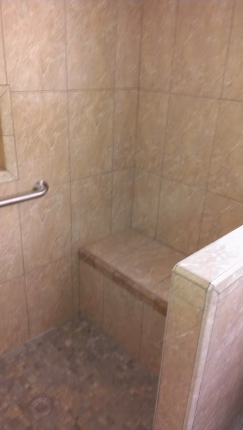 A tiled shower area with beige marble-patterned tiles covering the walls and a built-in bench. There is a metal grab bar mounted on the wall and a small recessed shelf for toiletries. The floor has small mosaic tiles with a drain visible.