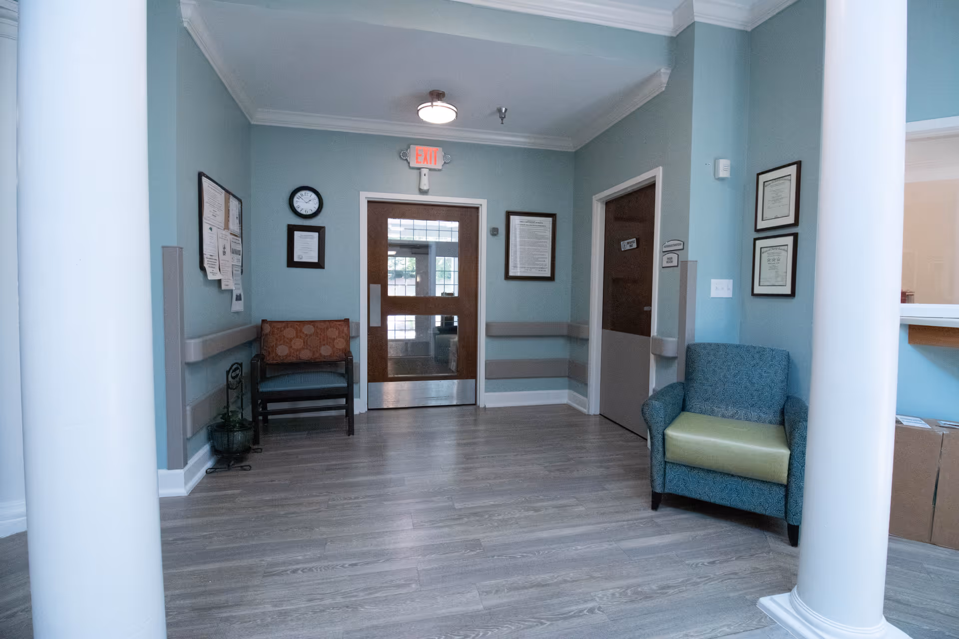 Small waiting area with chairs, white columns, wood-look flooring, framed notices, and double doors with an EXIT sign.