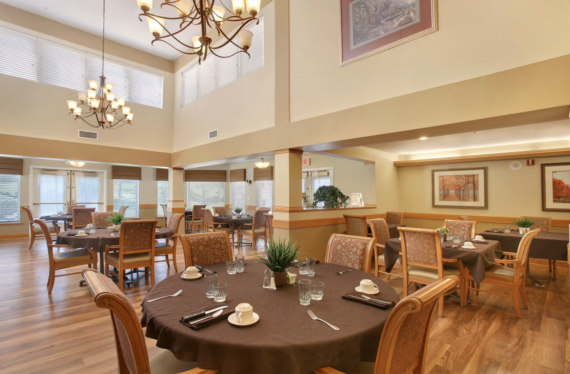 A spacious dining room with multiple round and rectangular tables covered with brown tablecloths, set with cups, glasses, and silverware. The room features wooden chairs with patterned upholstery, large windows with blinds, two chandeliers hanging from a high ceiling, and framed artwork on the walls.