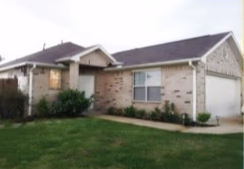Single-story brick house with a front lawn, a driveway leading to a garage, and some shrubs near the entrance.