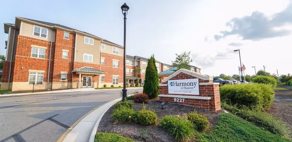 Exterior view of The Harmony Collection at Hanover Independent and Assisted Living facility showing a three-story brick and siding building, a landscaped area with bushes and flowers, a street lamp, and a sign with the facility name and address 8227.
