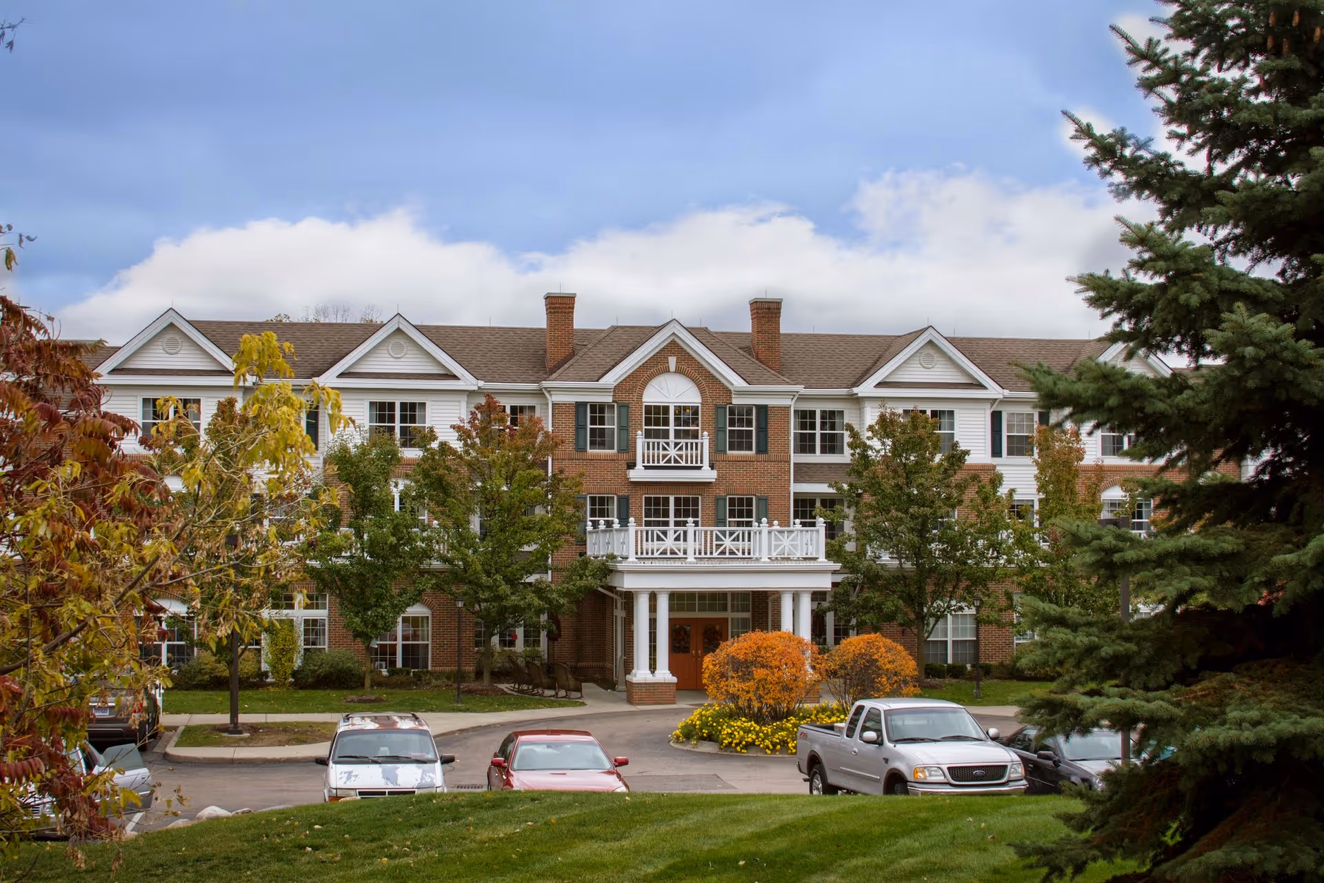 Front exterior view of a three-story senior living facility building with a brick and white siding facade, multiple windows, and a covered entrance with white columns. There are trees with autumn foliage and several parked cars in front of the building under a partly cloudy sky.