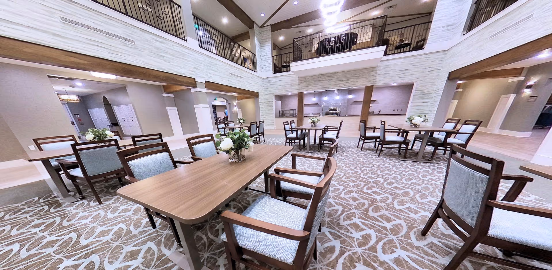 Spacious and well-lit dining area in a senior living facility with multiple wooden tables and chairs arranged on a patterned carpet. The room features a high ceiling with a second-floor balcony overlooking the space, and a kitchen or serving area is visible in the background. Floral centerpieces decorate the tables.