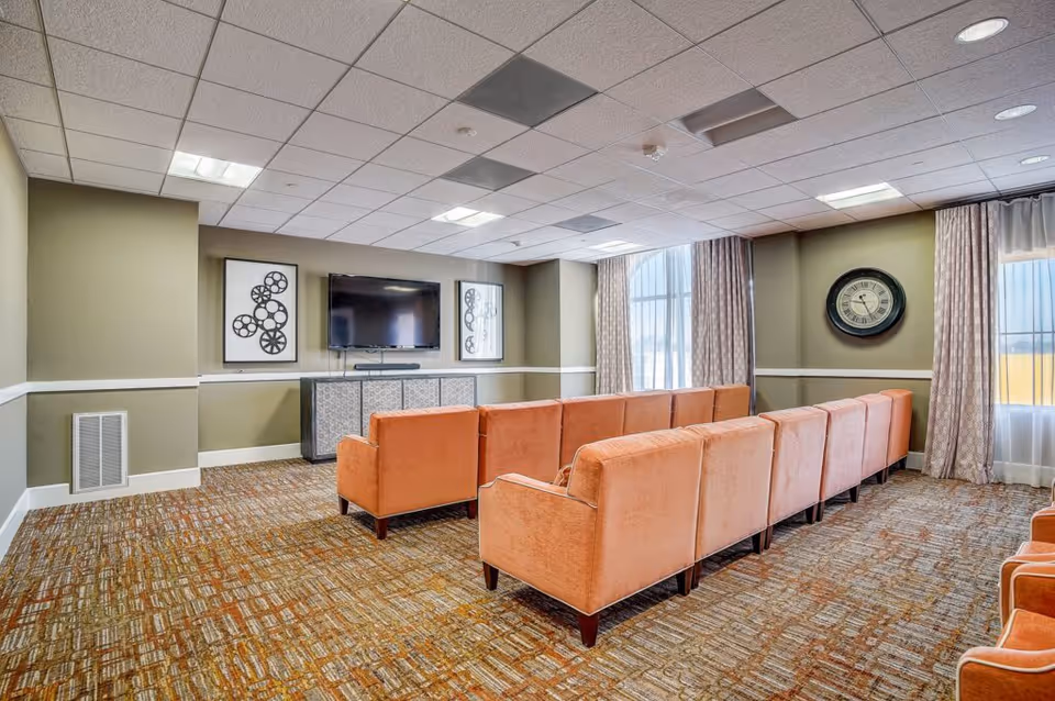 A cozy living room with two rows of orange upholstered sofas facing a wall-mounted flat screen TV. The room has patterned carpet flooring, beige walls with white trim, two large windows with curtains, a decorative wall clock, and framed artwork featuring film reels on either side of the TV.
