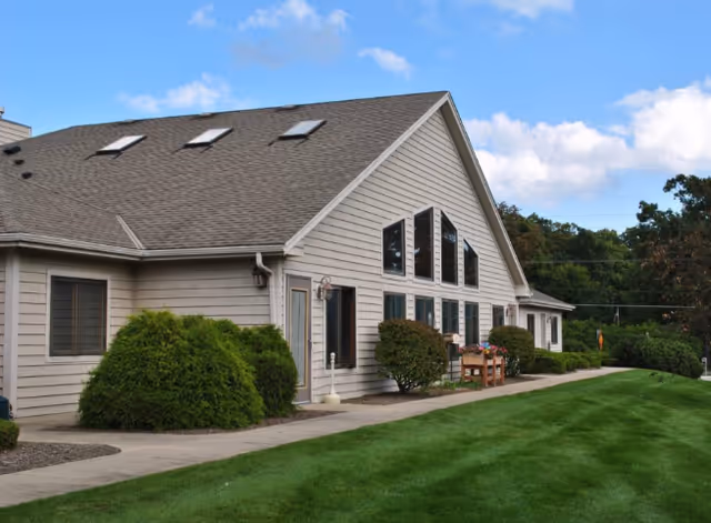 Exterior view of a single-story assisted living building with large gable windows, shrubs, a walkway, and a manicured lawn under a blue sky.