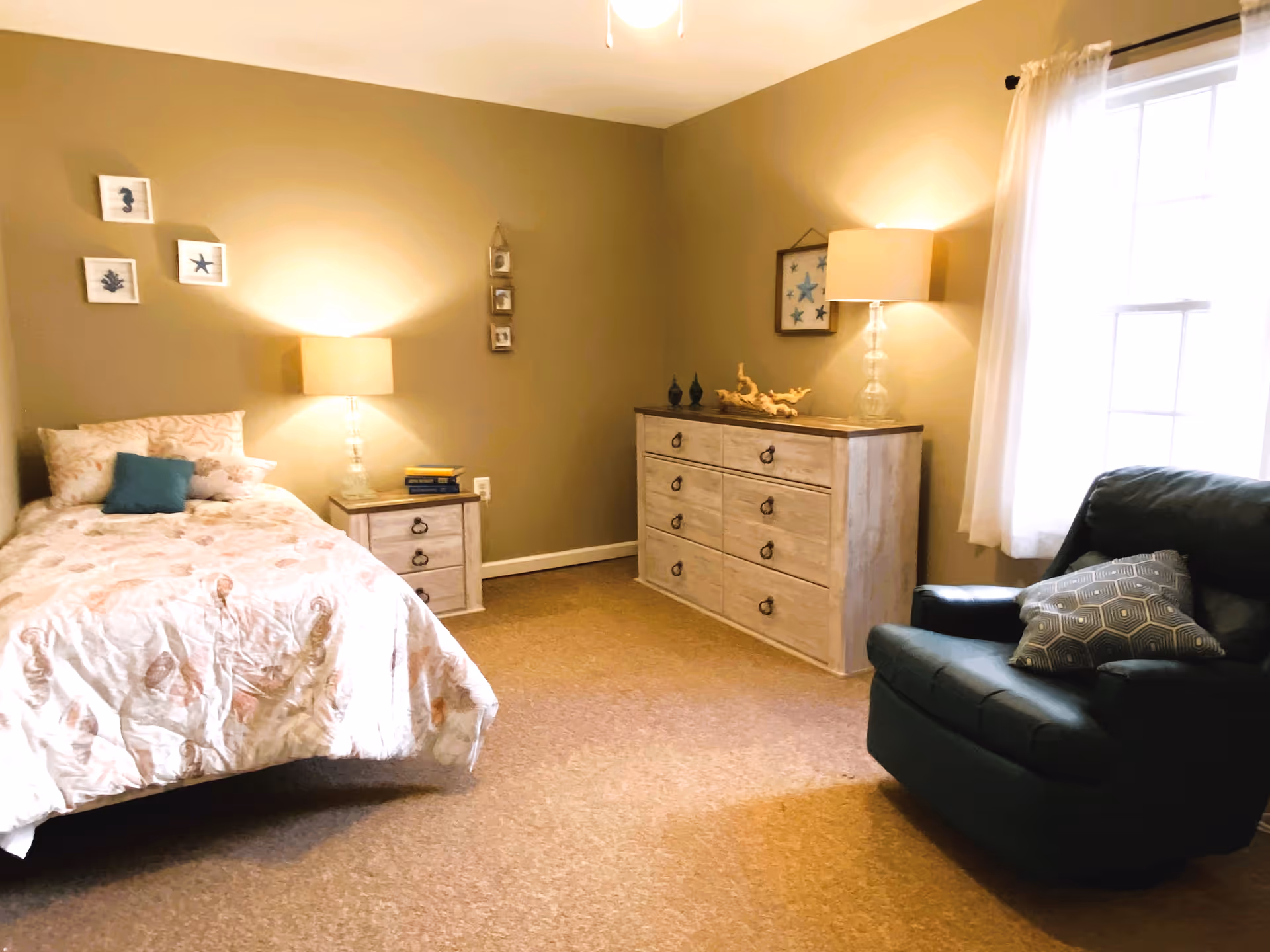 Cozy bedroom with a single bed, nightstand and lamp, a dresser with a lamp, and an armchair by a window.