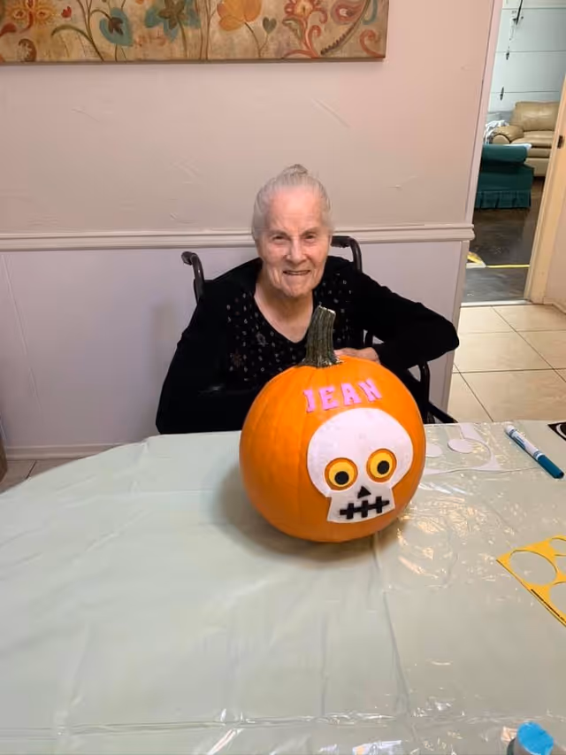 An elderly woman sitting in a wheelchair at a table with a decorated pumpkin in front of her. The pumpkin has a white skull face with yellow eyes and the name 'JEAN' written in pink letters above it. The room has light-colored walls and a floral painting hanging above the woman.