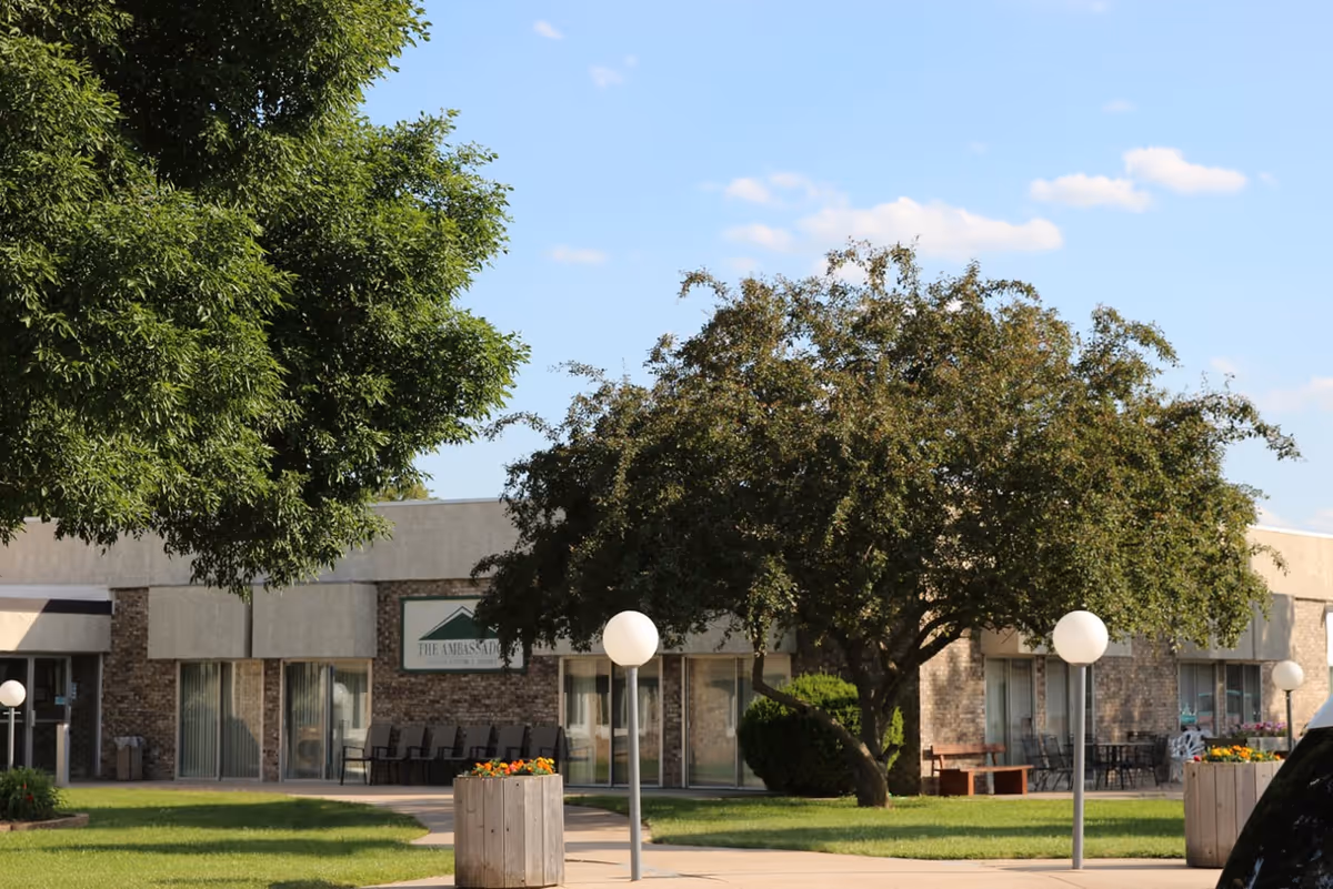 Exterior view of Ambassador Health of Sidney facility showing a single-story building with large windows, a tree in the foreground, green lawn, flower planters, and round white outdoor lights under a blue sky with some clouds.