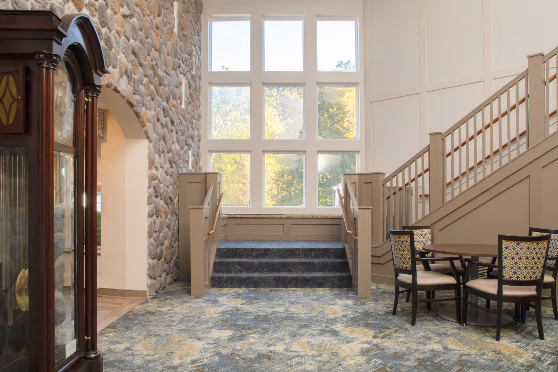 Interior view of a senior living facility featuring a stone accent wall, a large window with multiple panes letting in natural light, a staircase with beige railings, and a small seating area with a round table and four chairs with patterned upholstery. A wooden grandfather clock is visible on the left side.