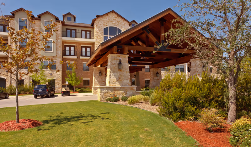 Exterior view of a multi-story assisted living facility with stone and brick facade, a large covered entrance with wooden beams, landscaped lawn with trees and shrubs, and a clear blue sky.