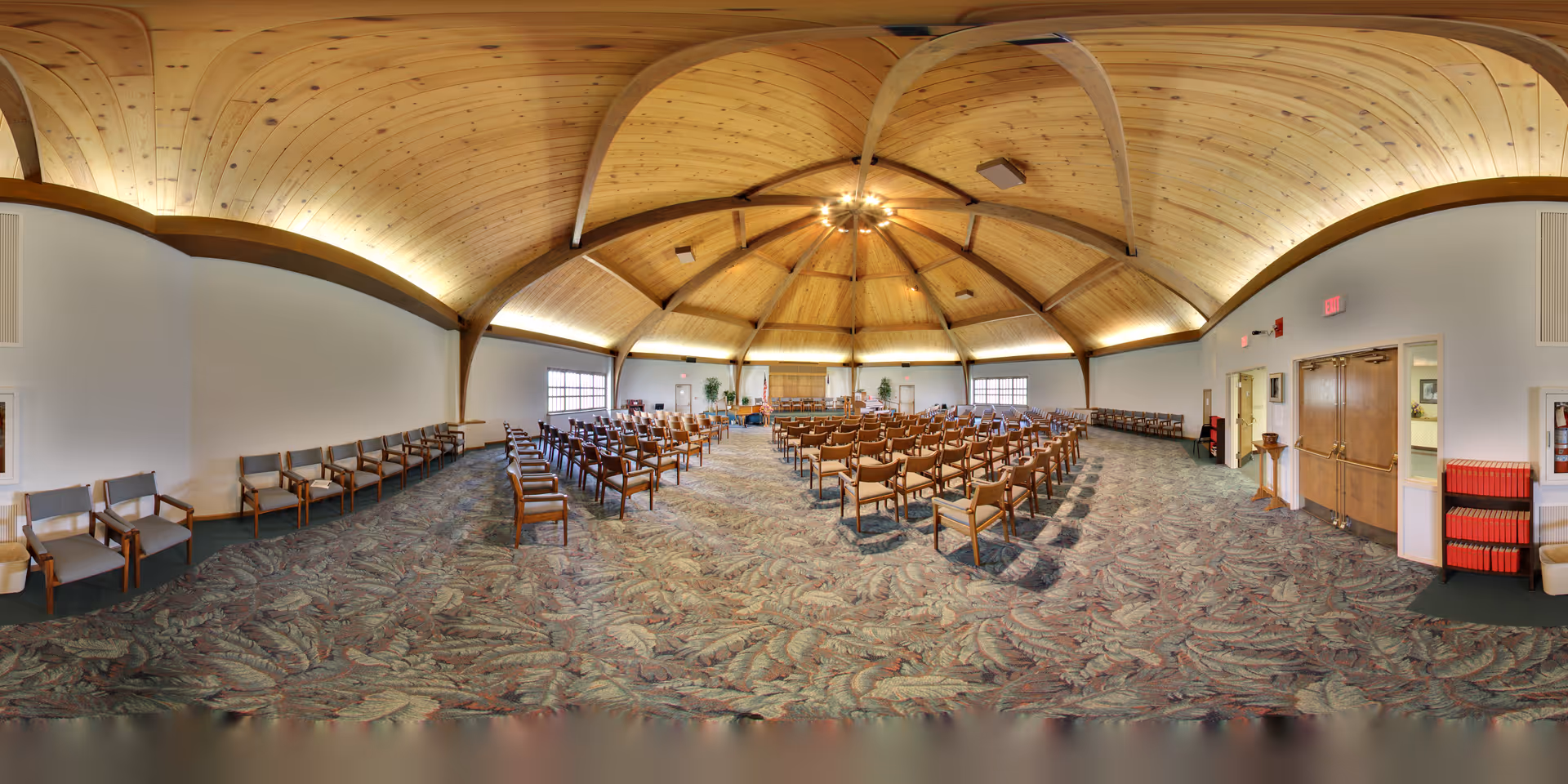 A large meeting or event room with rows of wooden chairs arranged facing a podium at the front. The room features a high, vaulted wooden ceiling with exposed beams and soft lighting. The floor is carpeted with a patterned design, and there are windows along the walls letting in natural light. The space appears clean and well-maintained, suitable for gatherings or presentations.