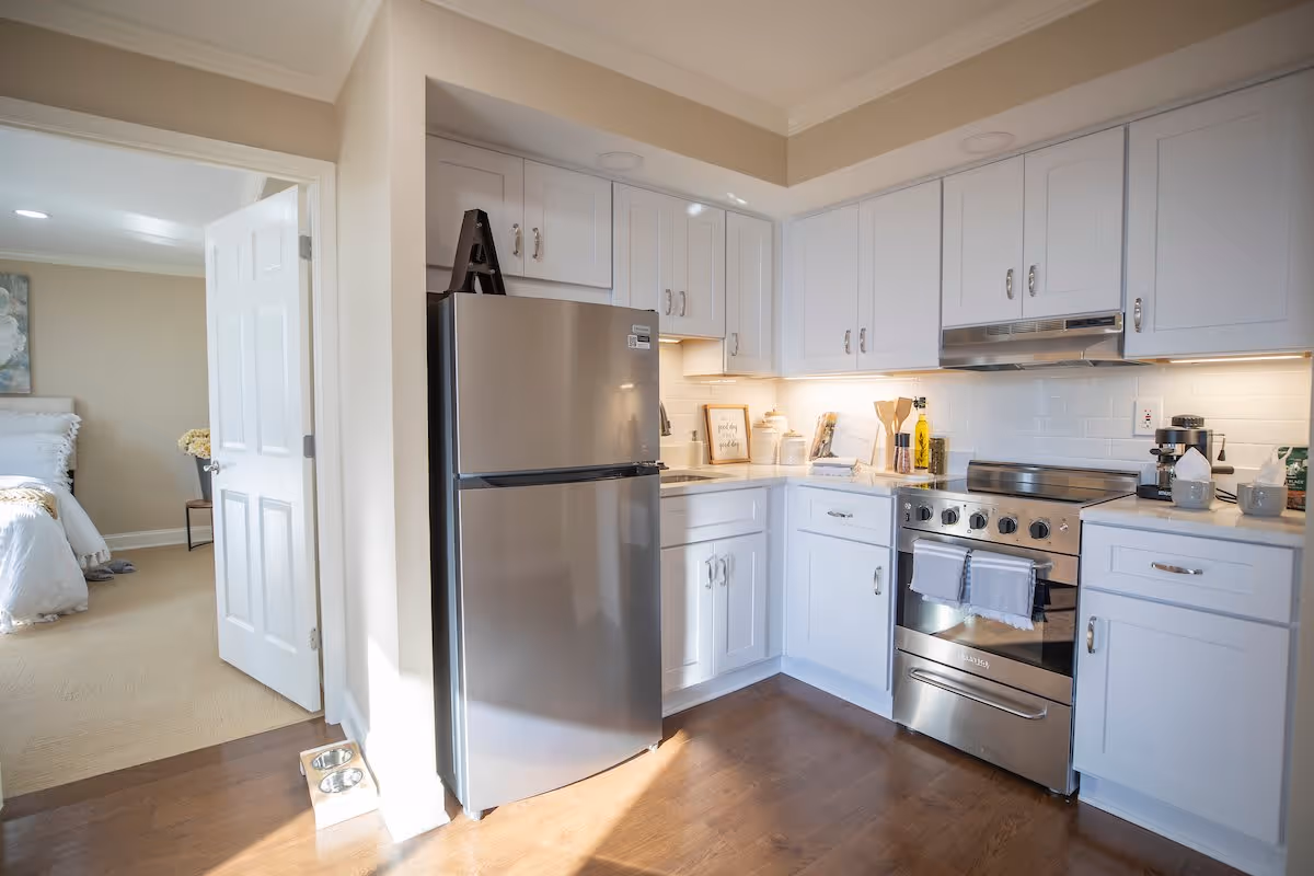 A bright kitchen with white cabinets, a stainless steel refrigerator, and a stainless steel stove with a range hood. The kitchen has a wooden floor and under-cabinet lighting. To the left, an open door leads to a bedroom with a bed and a nightstand with flowers.