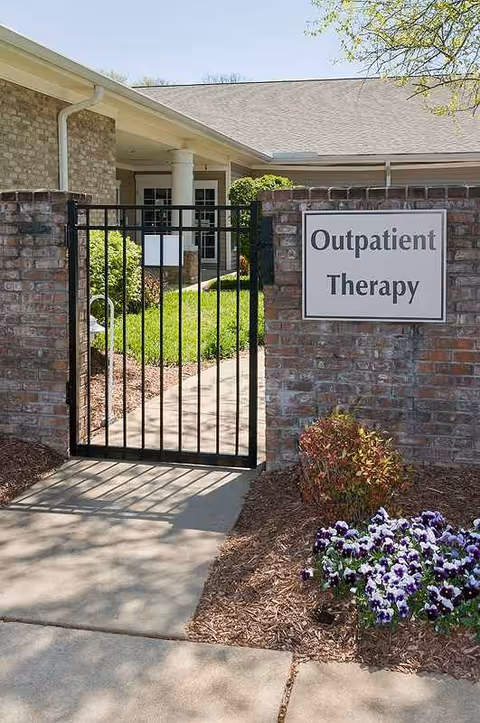 Gate leads to outpatient therapy entrance with brick wall and flowering landscaping.