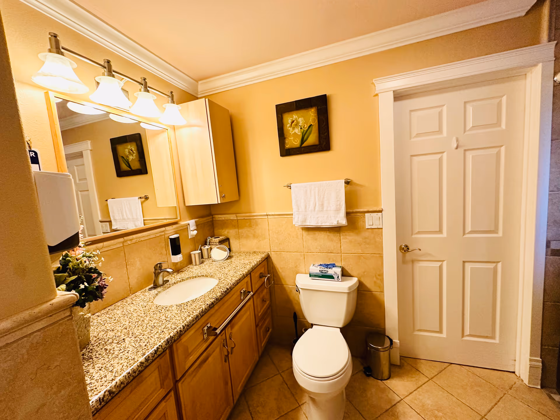 A clean and well-lit bathroom with beige tiled walls and floor. There is a granite countertop with a sink, wooden cabinets below, and a large mirror above. A white toilet is positioned next to the countertop with a box of tissues on top. A white towel hangs on a towel rack above the toilet, and a framed floral picture is mounted on the wall. A closed white door is visible on the right side.